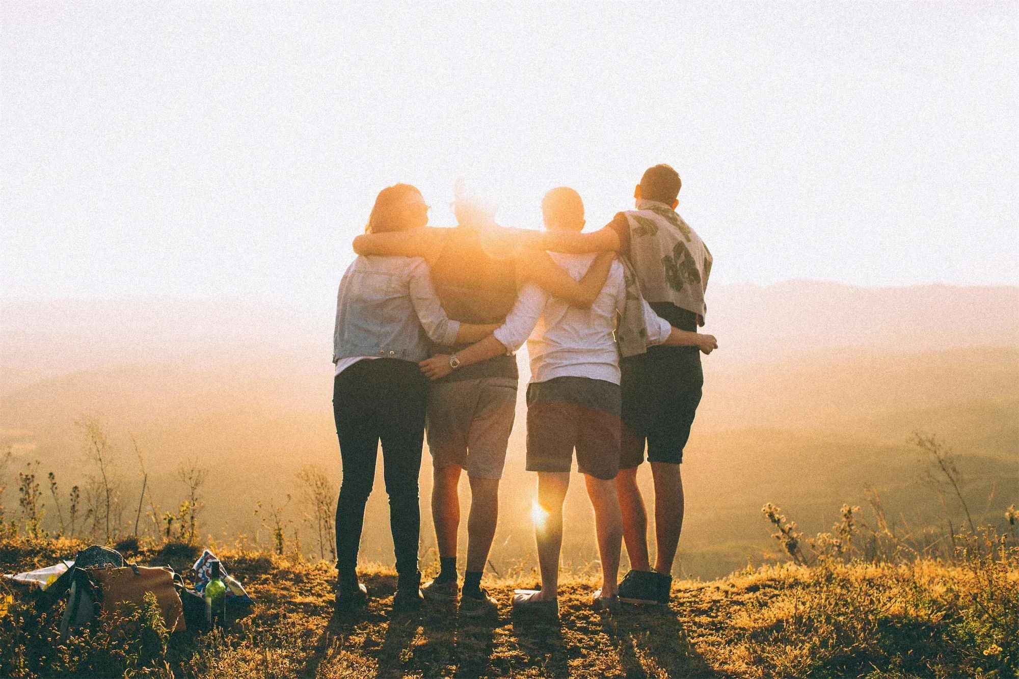 Four friends huddle together at sunset in grassy field, backlit by golden light