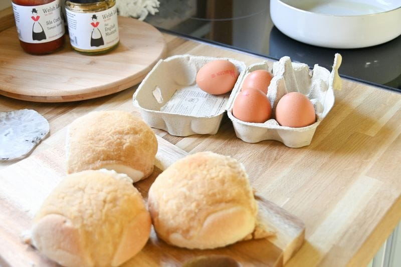 Fresh bread rolls and brown eggs in carton on wooden cutting board with kitchen background